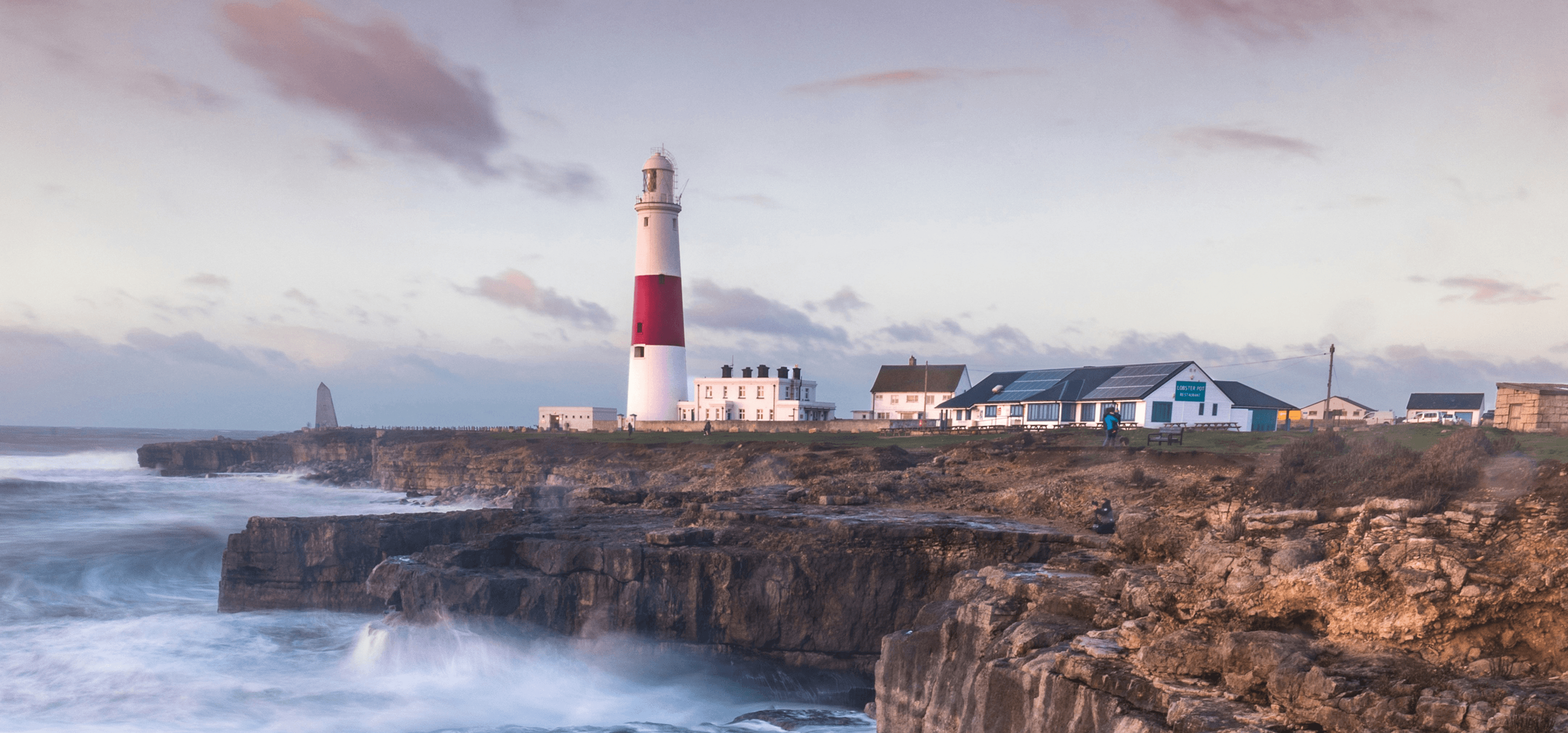 Las olas rompen en un acantilado británico con un faro en la distancia.